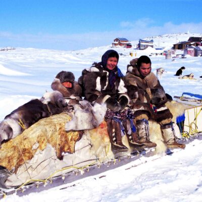 An Inuit woman, teenager, and child sit atop a sled on a snow-covered field
