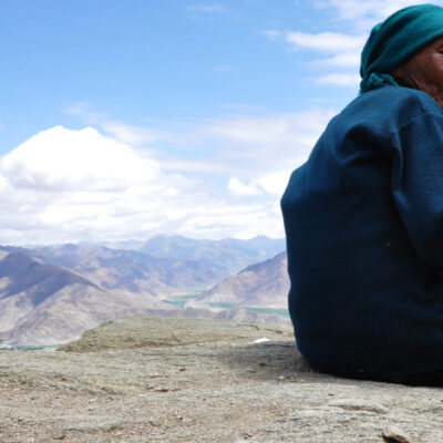 Photo of a Tibetan woman, face turned toward the camera, sitting and overlooking a mountain range.