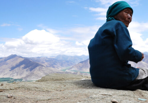 Photo of a Tibetan woman, face turned toward the camera, sitting and overlooking a mountain range. 